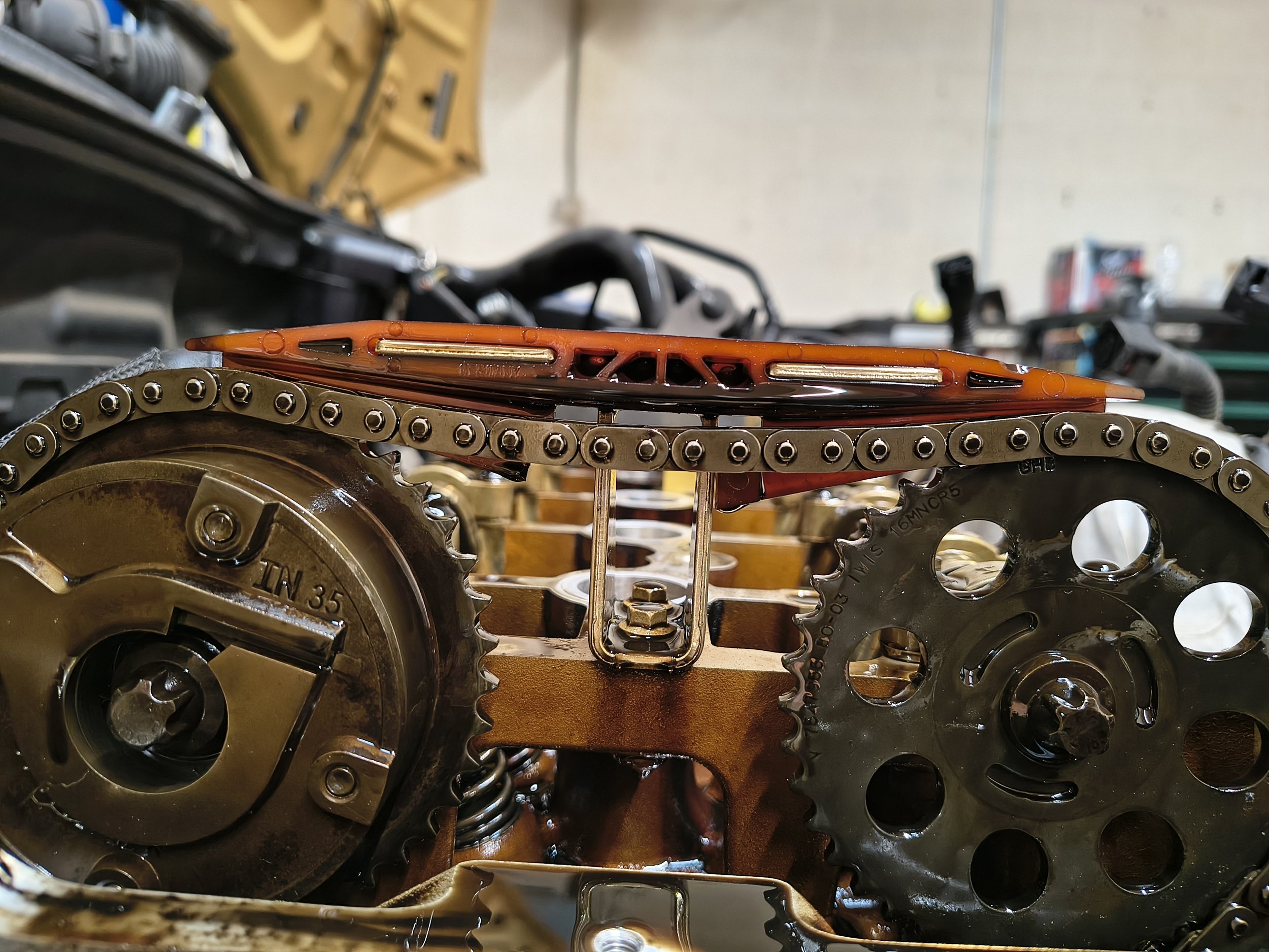 Close-up of interconnected metal gears and chains inside a mechanical device, with visible oil or lubricant and a blurred workshop background.