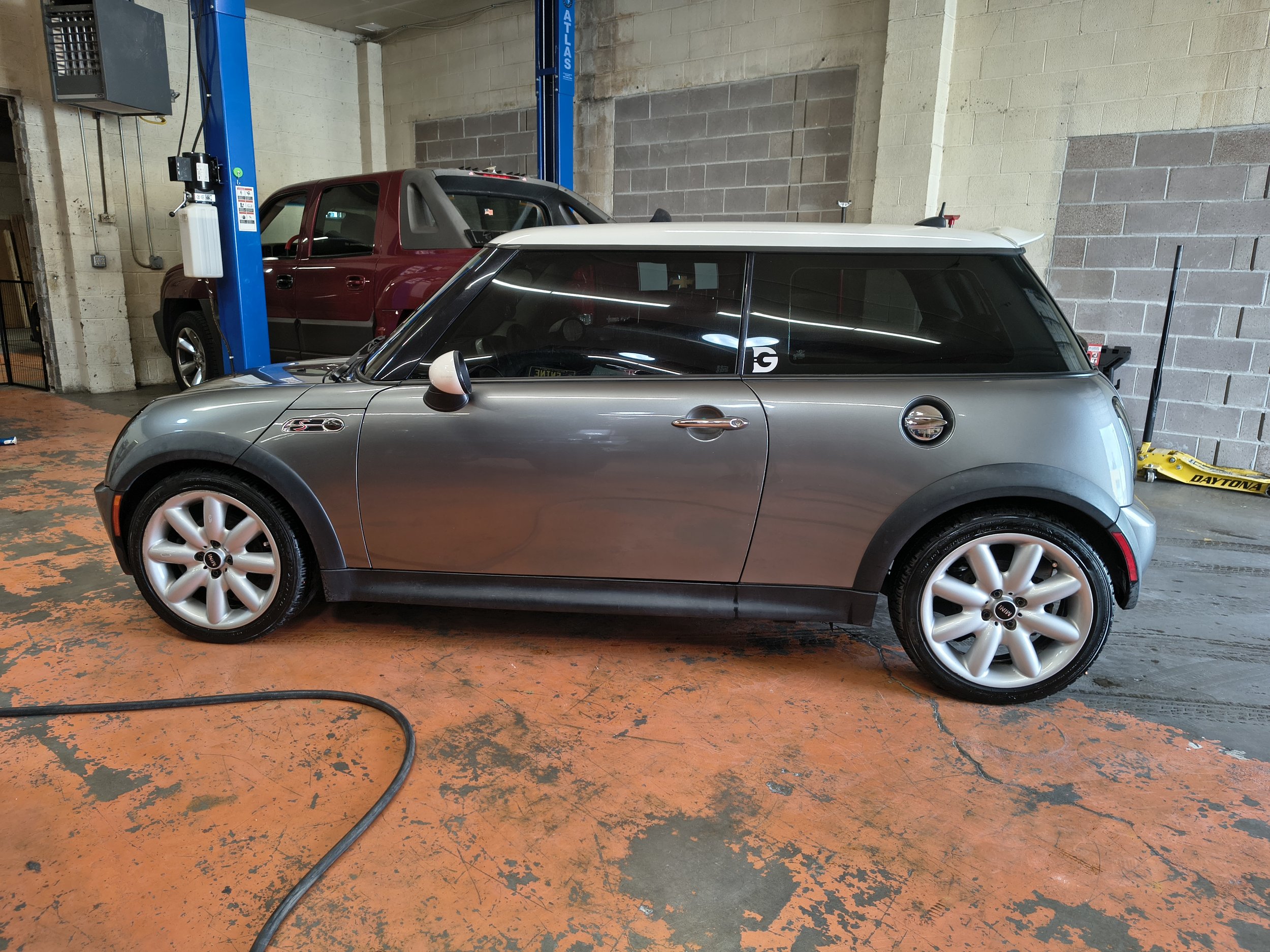 A silver Mini Cooper with a black roof parked inside a garage next to a red pickup truck on a blue hydraulic lift. The garage has a concrete block wall and an orange and gray concrete floor.