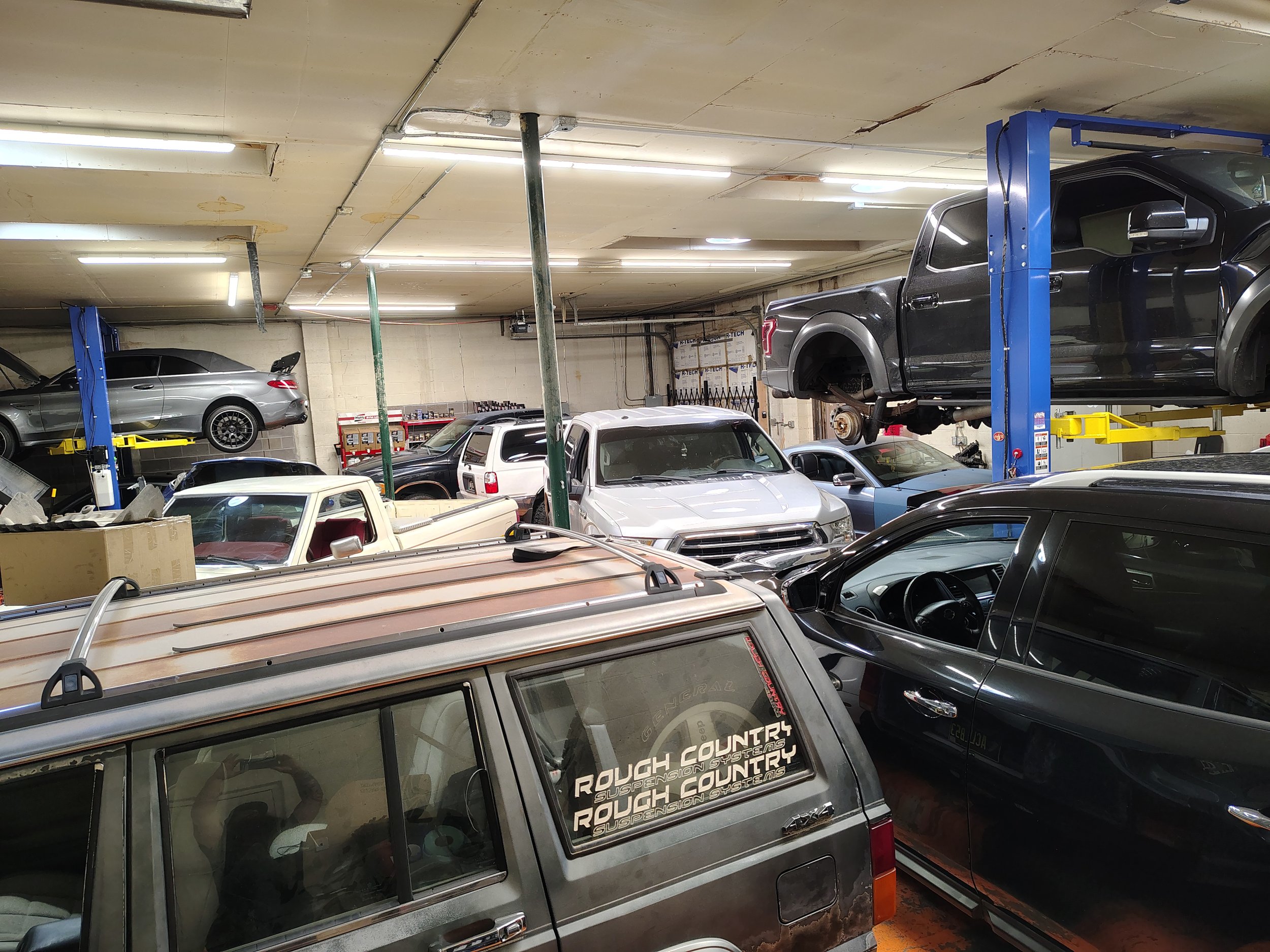 Multiple vehicles parked in a basement garage, including a black pickup truck on a lift, a silver car on the left, and a van with stickers reading 'Rough Country Suspension Systems' in the foreground.