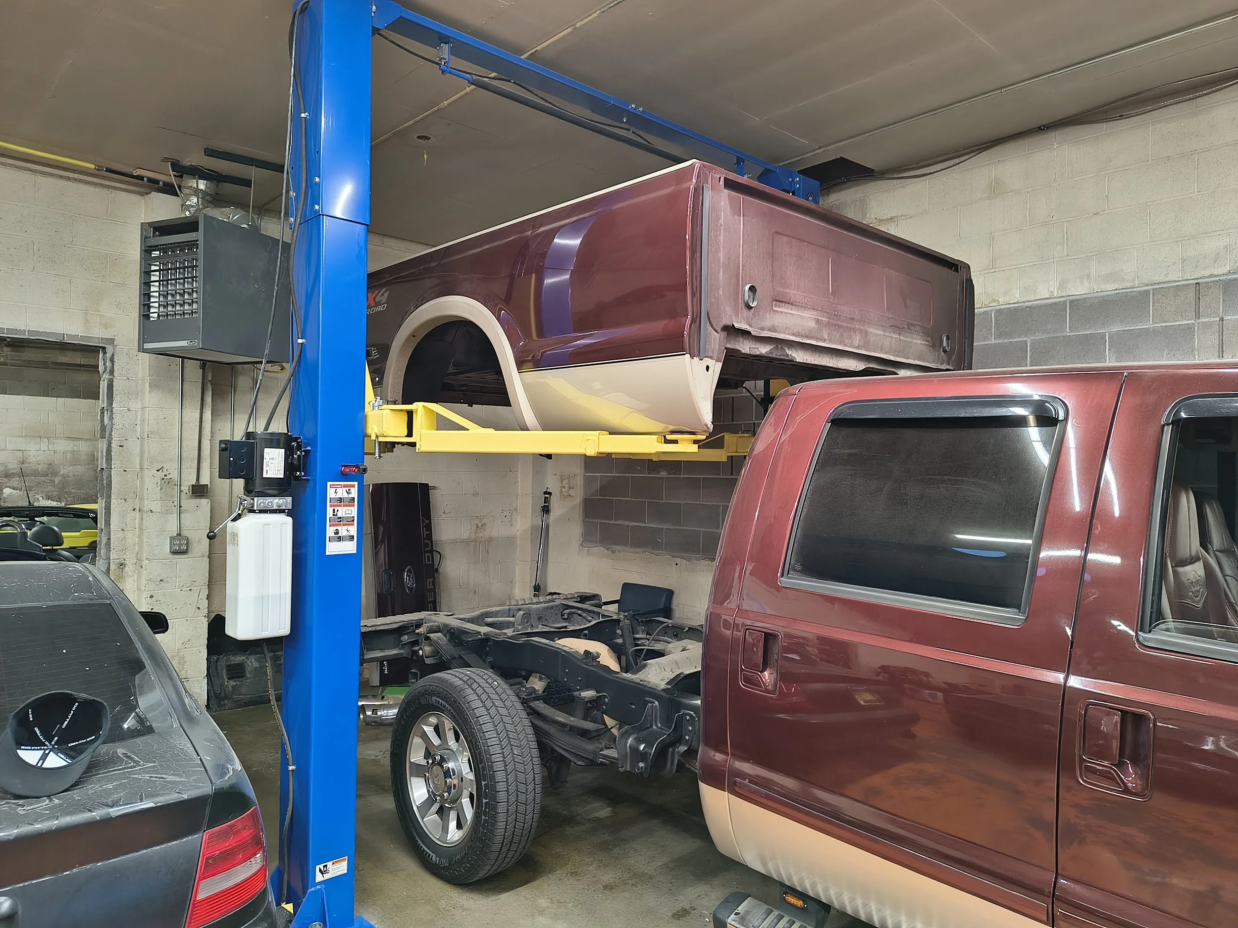 A vehicle repair shop with a blue hydraulic lift holding up a maroon pickup truck, which is being serviced without its rear wheels. Other cars are visible, including a silver car in the foreground and a red vehicle on the right.