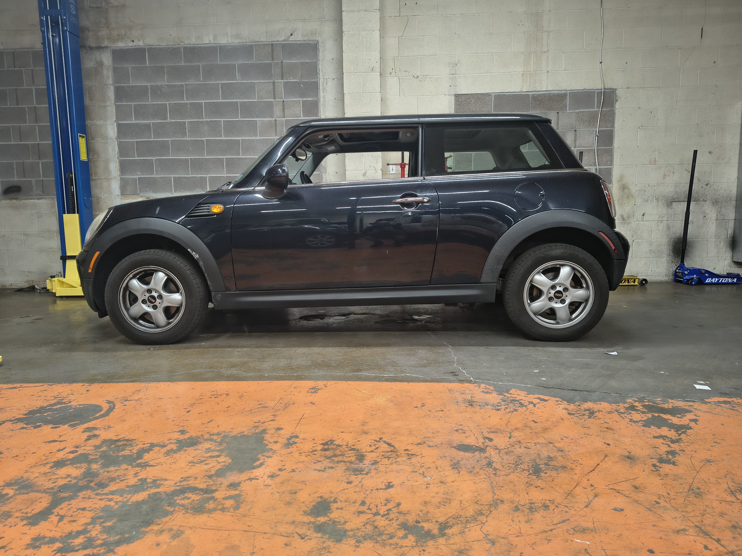 A black Mini Cooper car is parked inside a garage or workshop. The car is positioned next to a blue hydraulic lift on the left and a black jack on the right. The background features a gray cinder block wall with some exposed wiring.