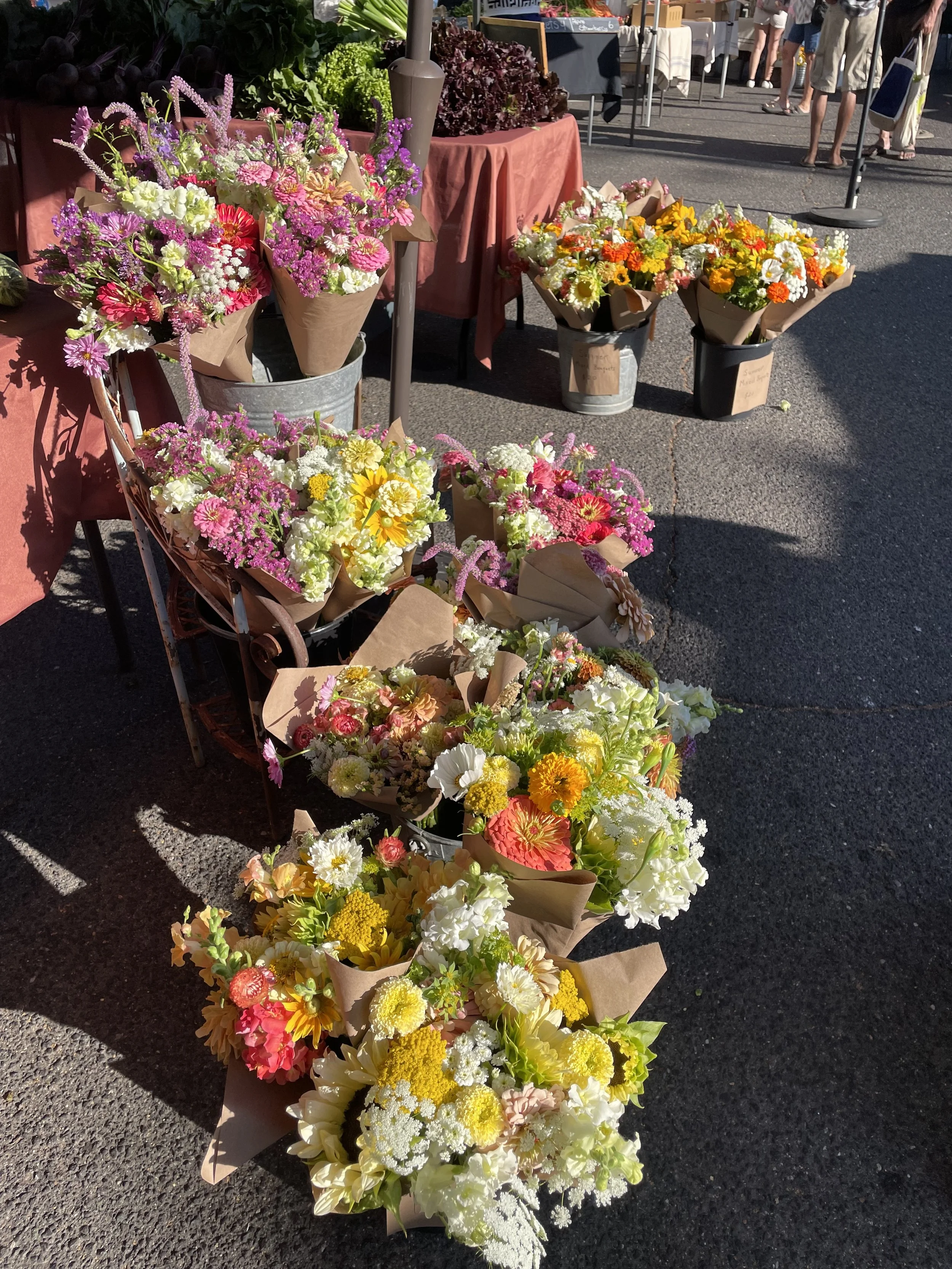 Various colorful flower bouquets displayed at the Sonora Farmers Market, with people walking in the background.