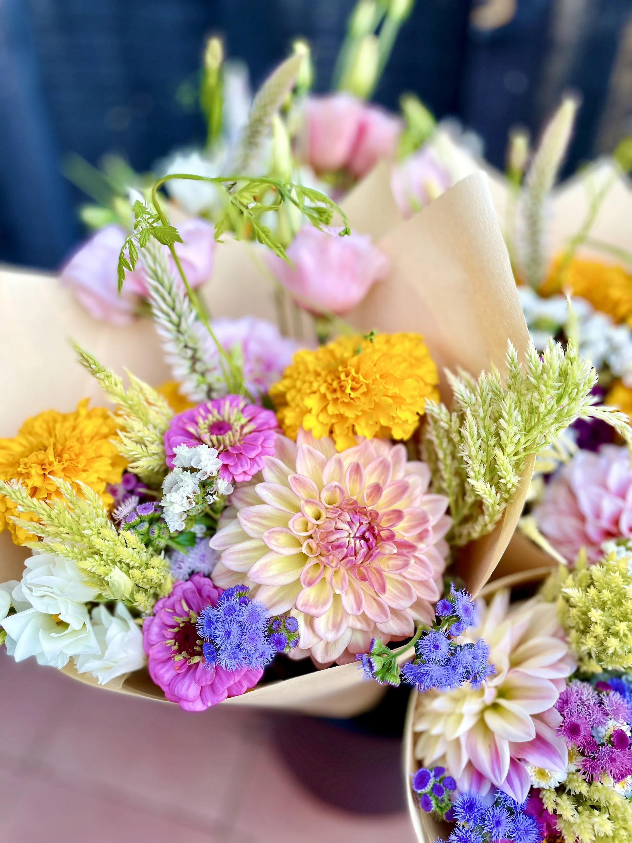 Colorful bouquet of various flowers including pink, yellow, white, purple, and lavender blooms wrapped in light brown paper.