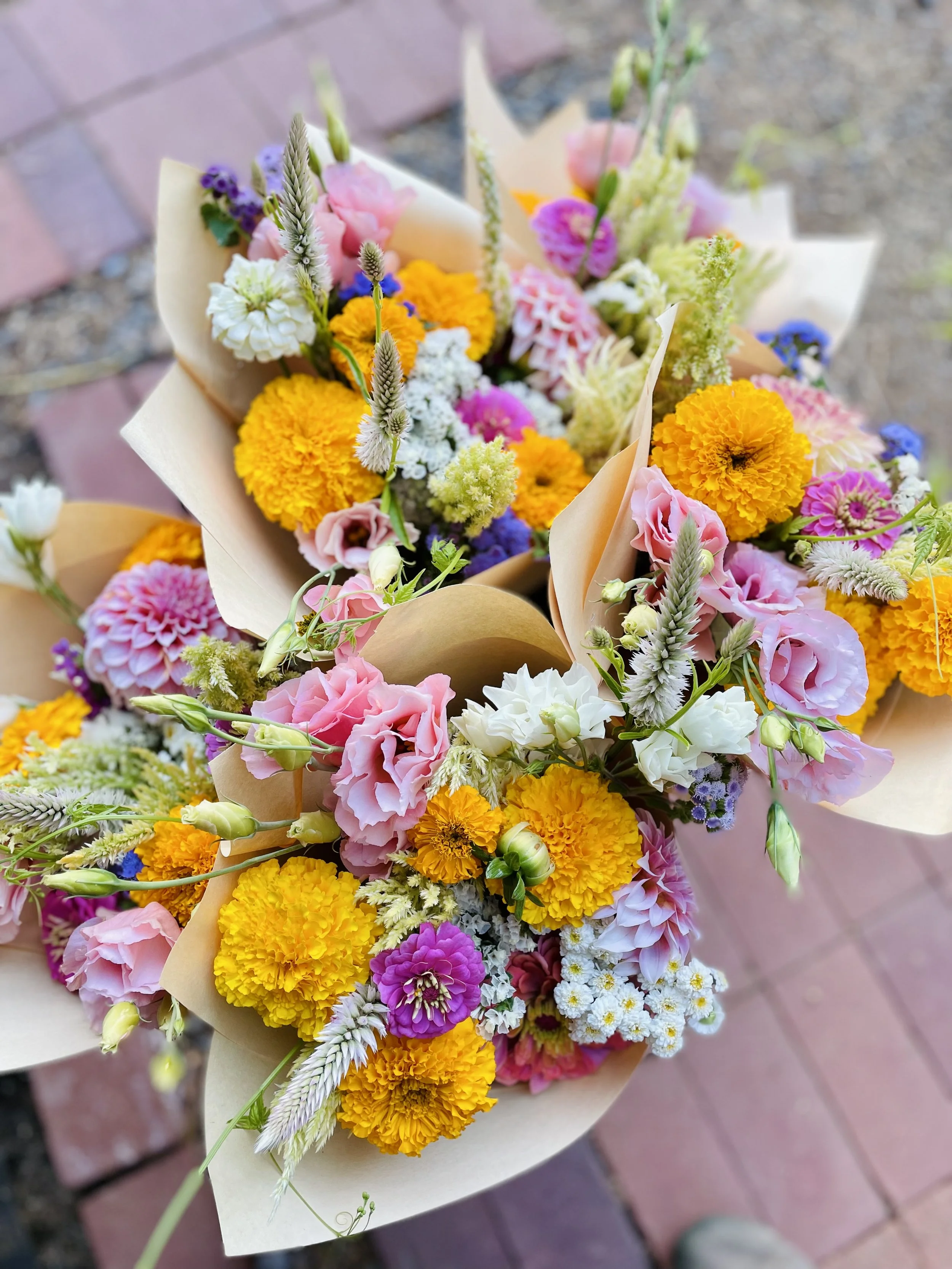 Colorful flower bouquets wrapped in brown paper, featuring pink, yellow, purple, white, and peach blossoms, on a brick and dirt background.