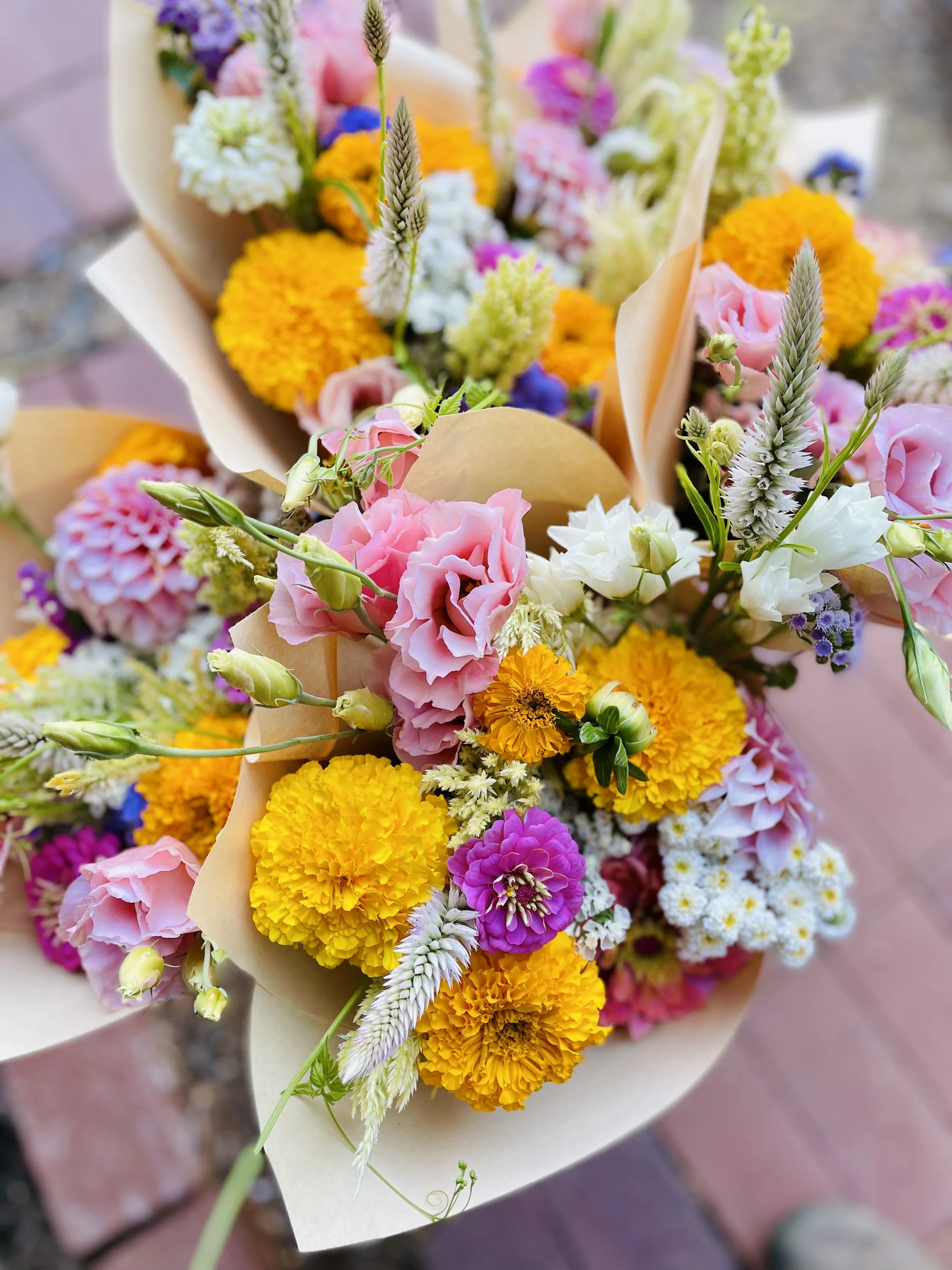 Colorful mixed flower bouquet including pink lisianthus, yellow marigolds, purple zinnias, white astilbe, and other assorted flowers wrapped in beige paper.