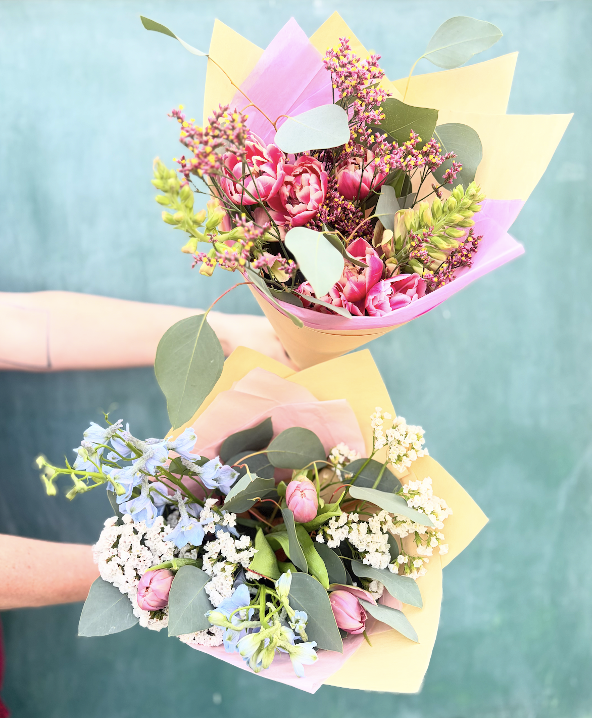 Two bouquets of flowers held by a person against a teal background. The bouquets include pink tulips, white blossoms, light purple flowers, and green leaves, wrapped in pastel-colored paper.