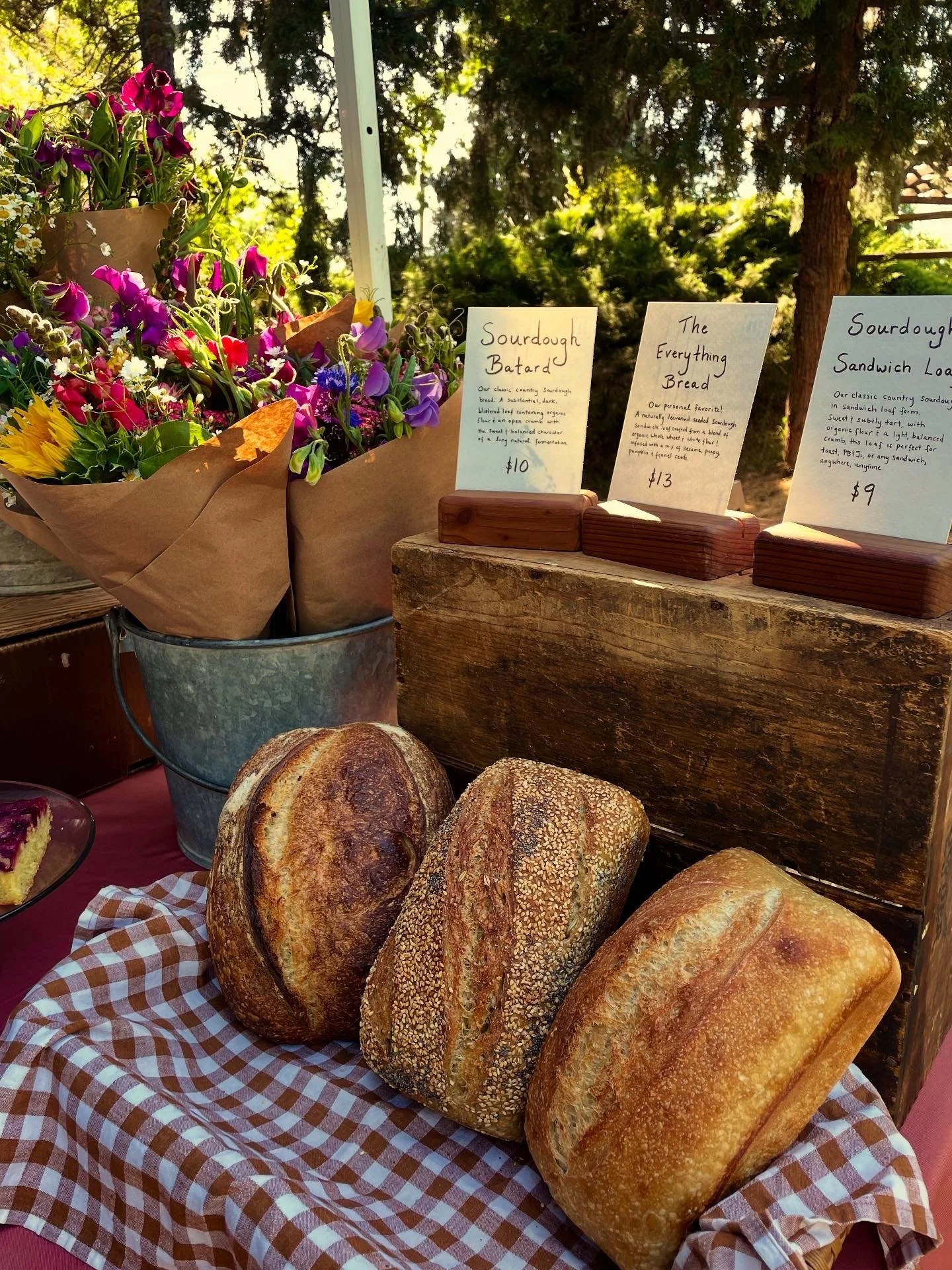 Assorted loaves of bread on a checkered cloth, with a metal bucket of flowers and handwritten signs describing sourdough bread and sandwiches at the Murphys Farmers Market.