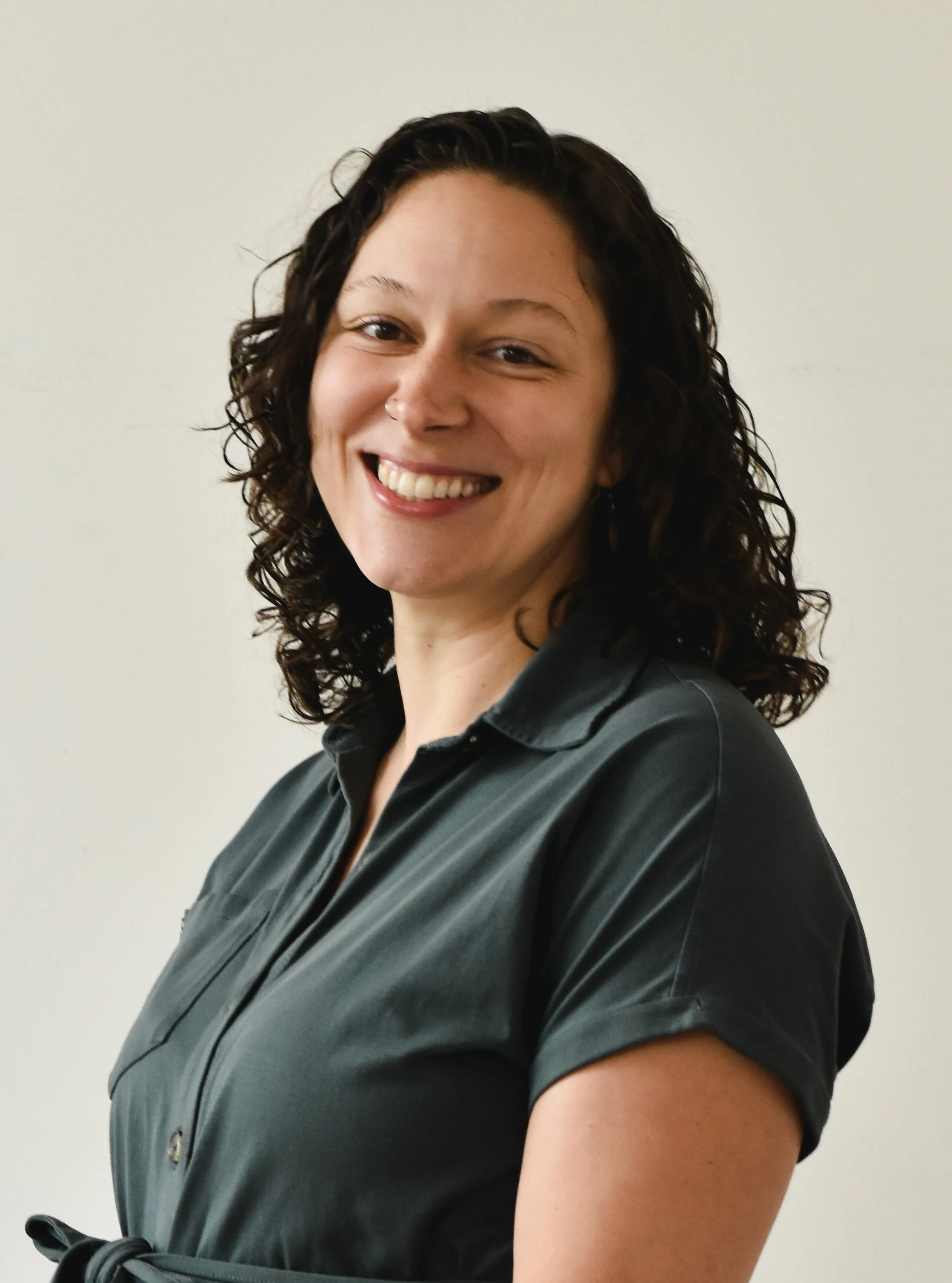 A woman with curly dark hair smiling, wearing a dark gray collared shirt, standing against a plain light-colored wall.