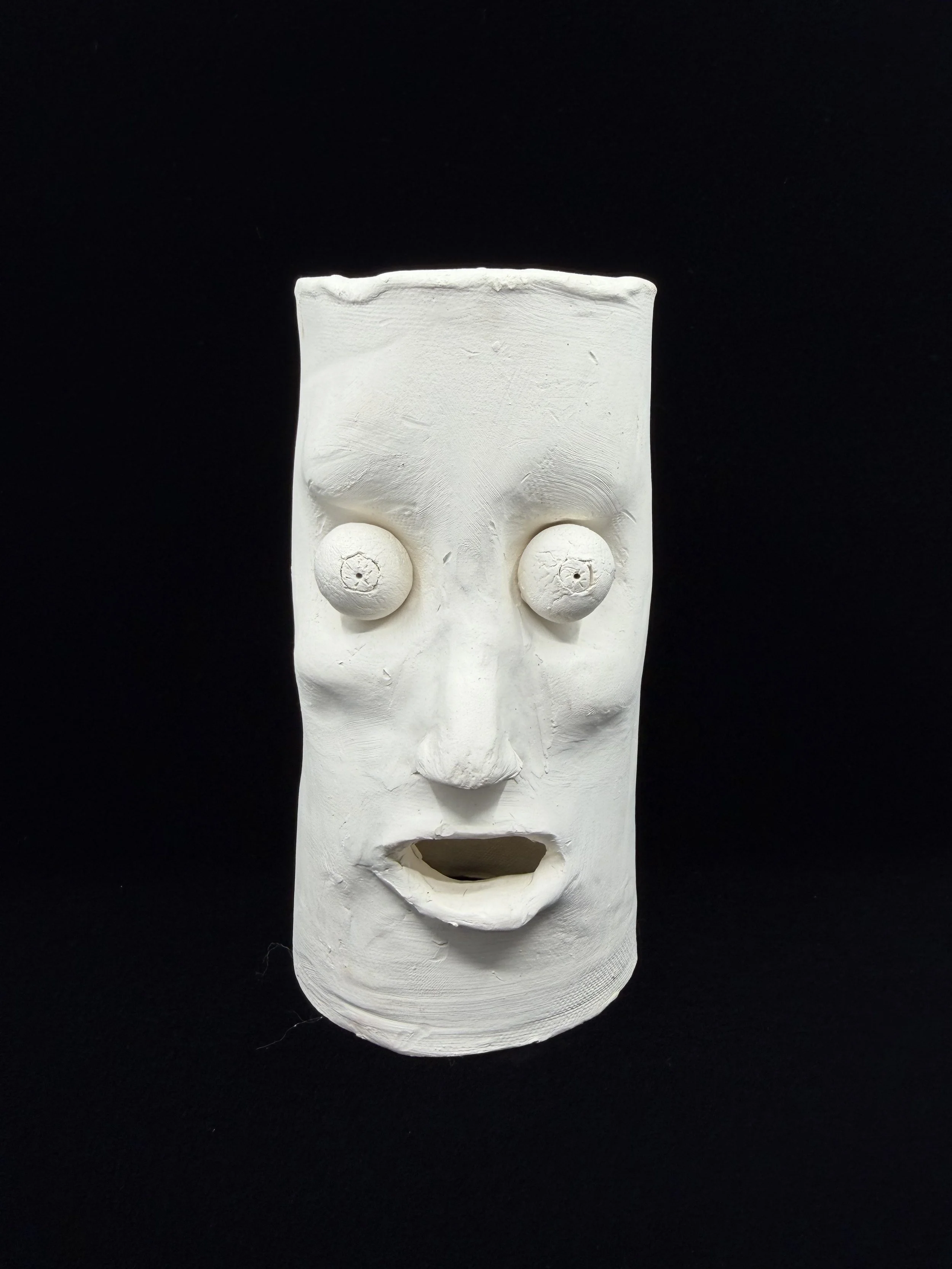 White tribal-style clay sculpture with exaggerated facial features and hollow eyes, against a black background.
