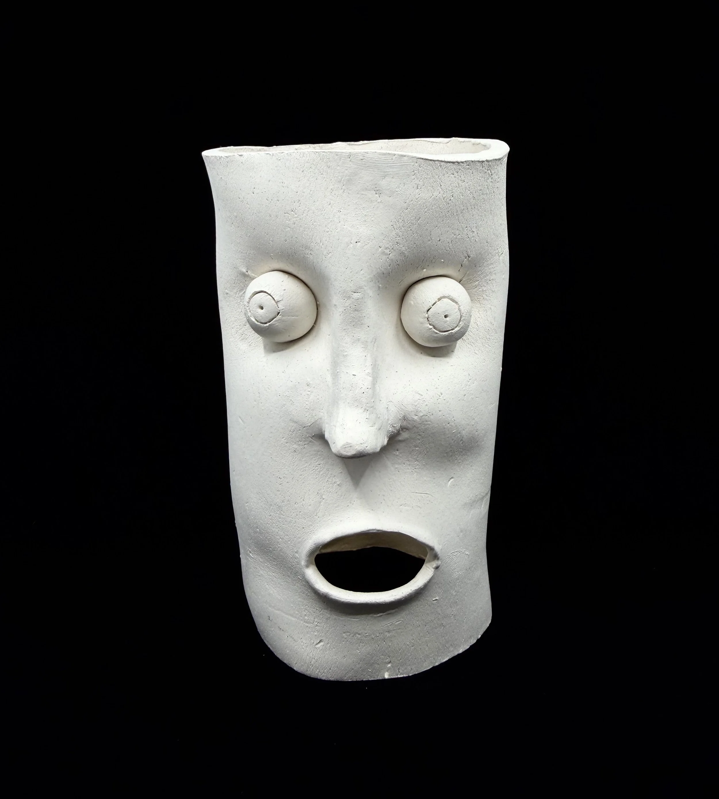 White ceramic figure resembling a human face with exaggerated features, including round eyes, a prominent nose, and an open mouth, set against a black background.