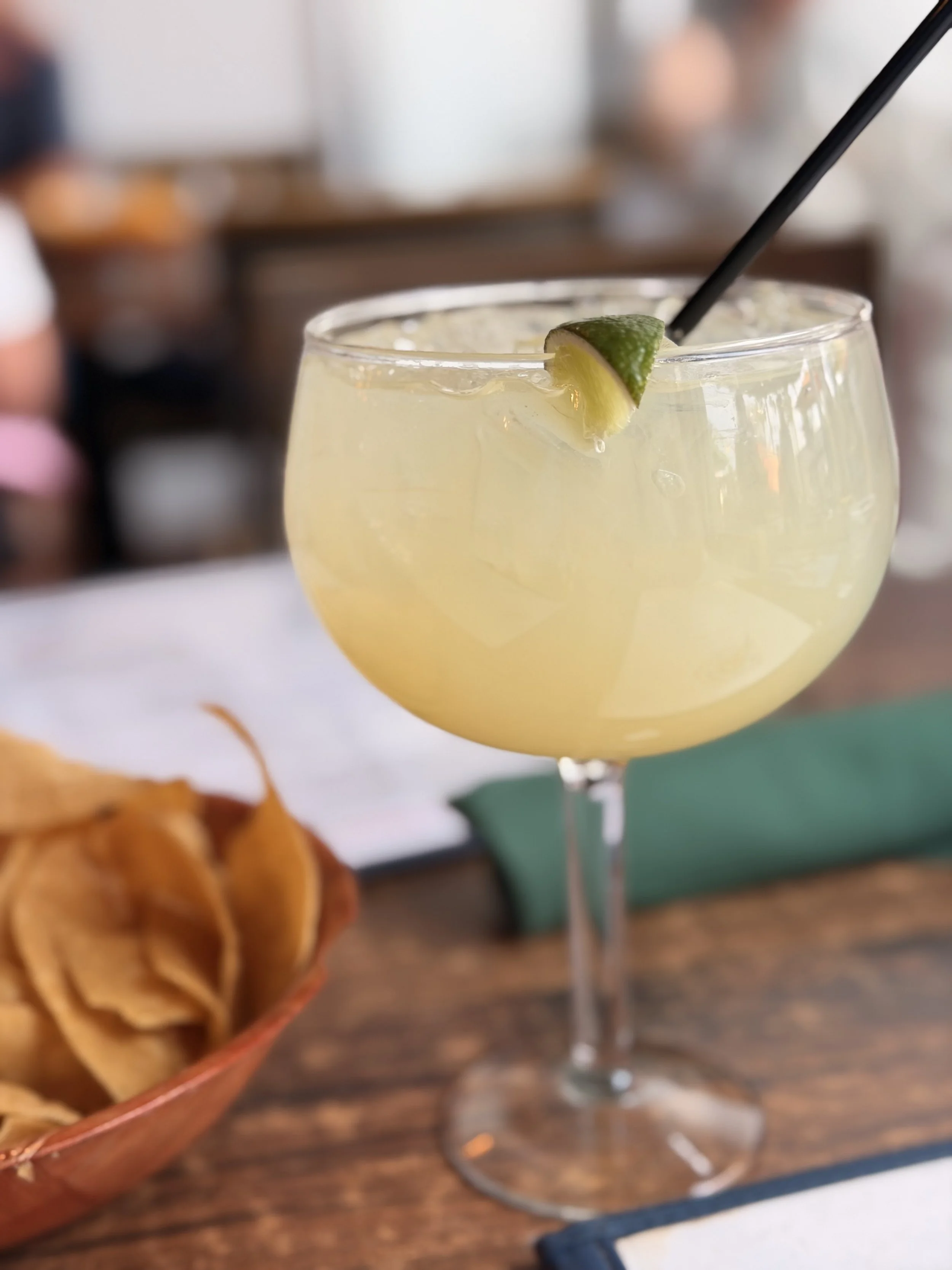 Close-up of a yellow cocktail with a lime wedge garnish in a wide glass, with a straw, on a wooden table with a blurred background.
