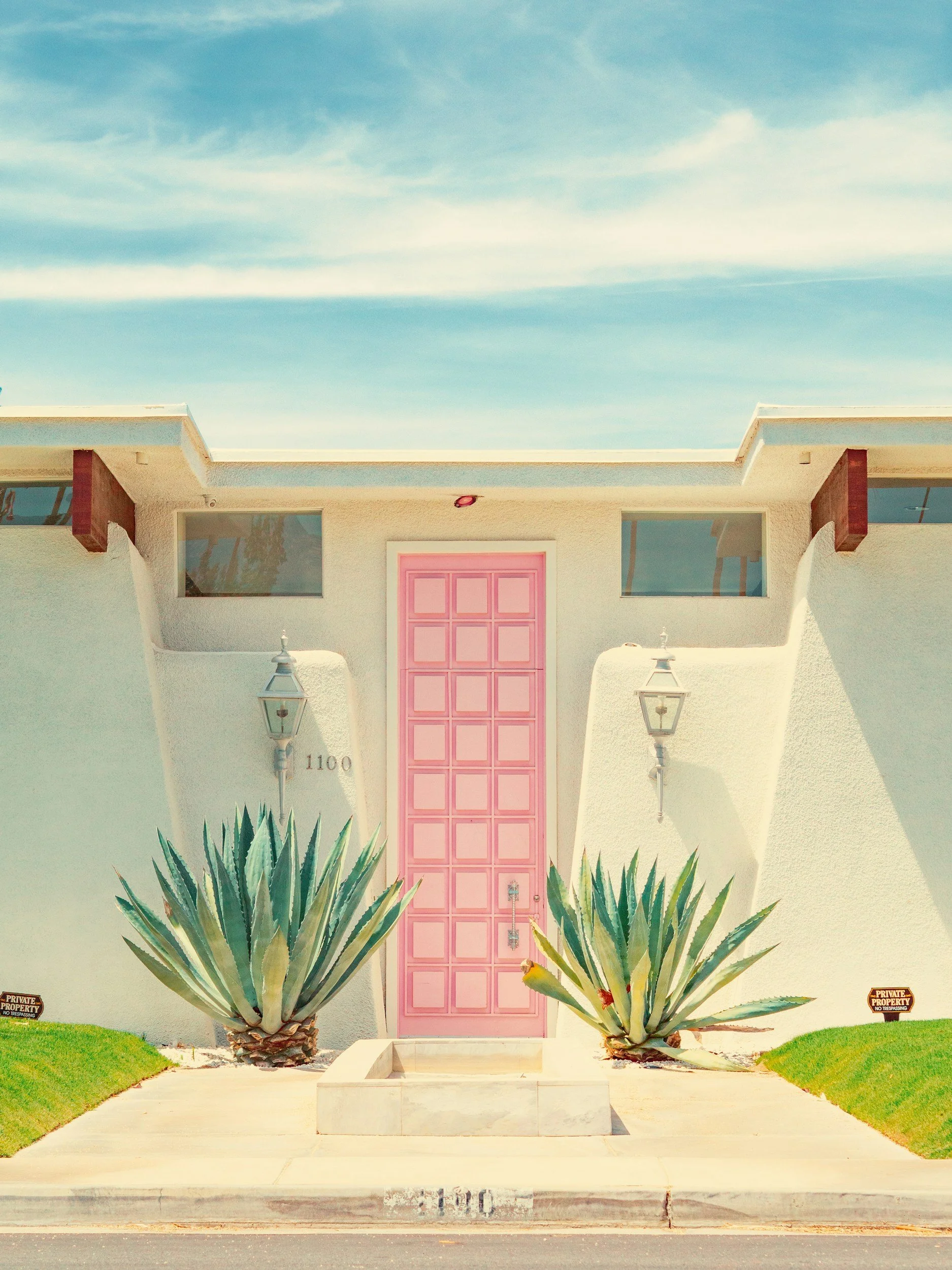 A modern house with a pink door, white textured exterior walls, and two large agave plants in the front yard. There are signs indicating private property on either side of the plants. The house has a flat roof with wooden accents underneath the roof overhang and two large windows.