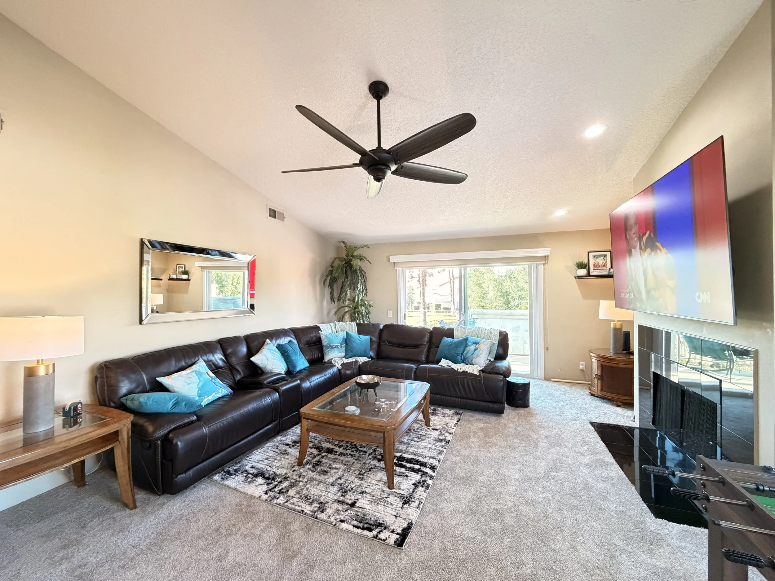 Living room with a black leather sectional sofa decorated with blue pillows, a wooden coffee table, a large wall-mounted TV, a fireplace, a side table with a lamp, and a ceiling fan. There are two windows allowing natural light and a mirror on the wa
