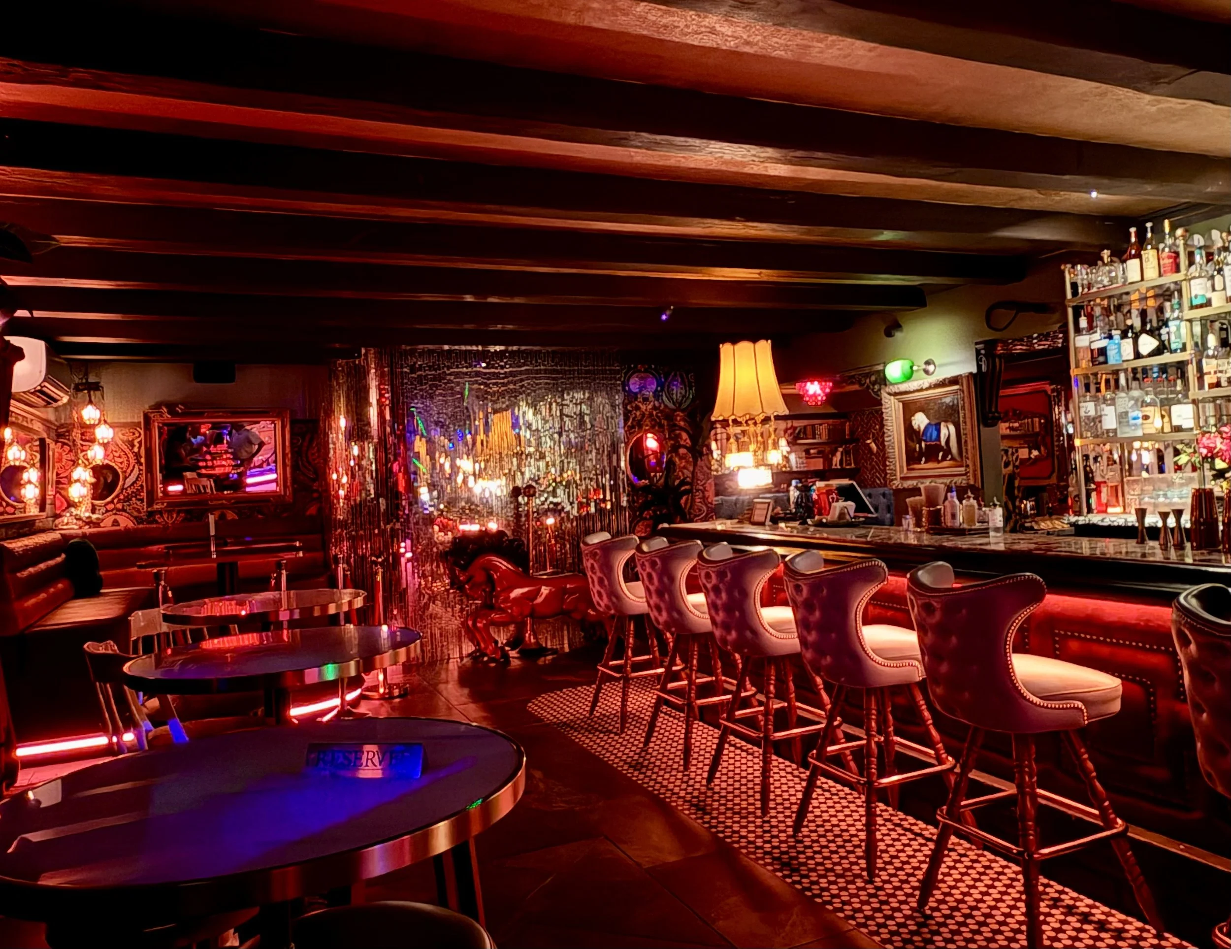 Interior of a dimly lit bar or lounge with red and warm lighting, a bar counter with bottles on shelves, and seating arrangements including bar stools and chairs.