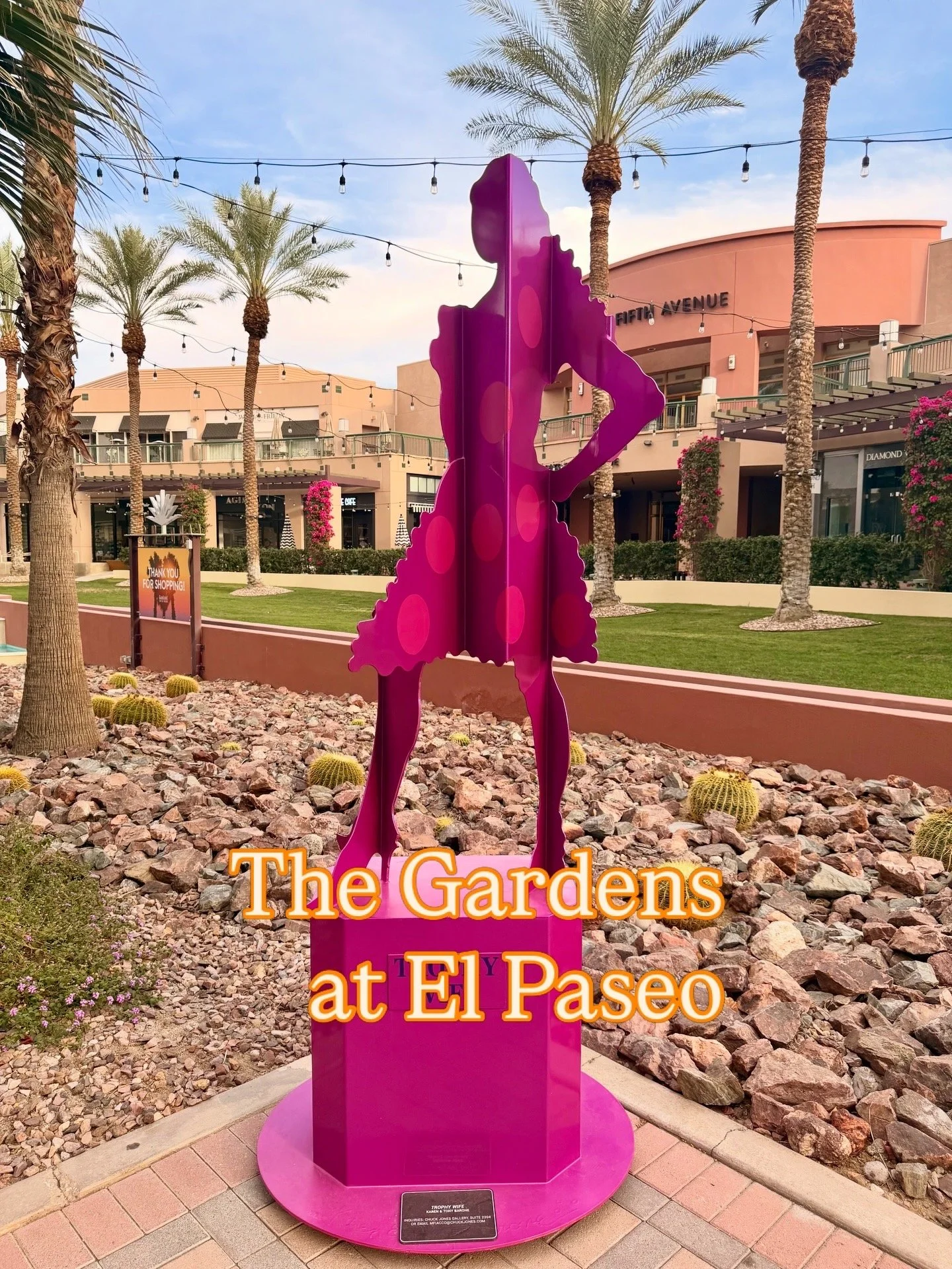 Pink decorative sculpture of a woman wearing a dress and a bow in her hair, standing in an outdoor shopping center with palm trees, buildings, and string lights. The text on the sculpture reads "The Gardens at El Paseo."