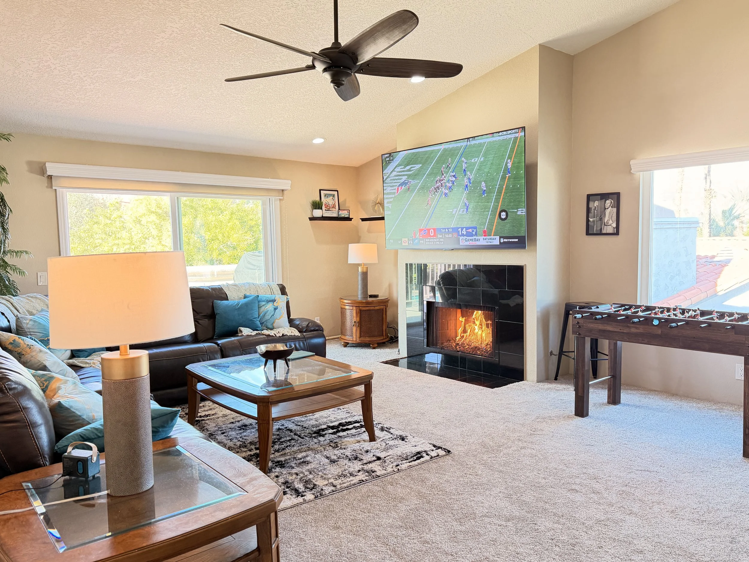 A living room with a ceiling fan, a large wall-mounted television showing a football game, a fireplace with flames, a sectional sofa with blue and patterned pillows, a wooden coffee table, a lamp, a foosball table, and large windows allowing natural light.