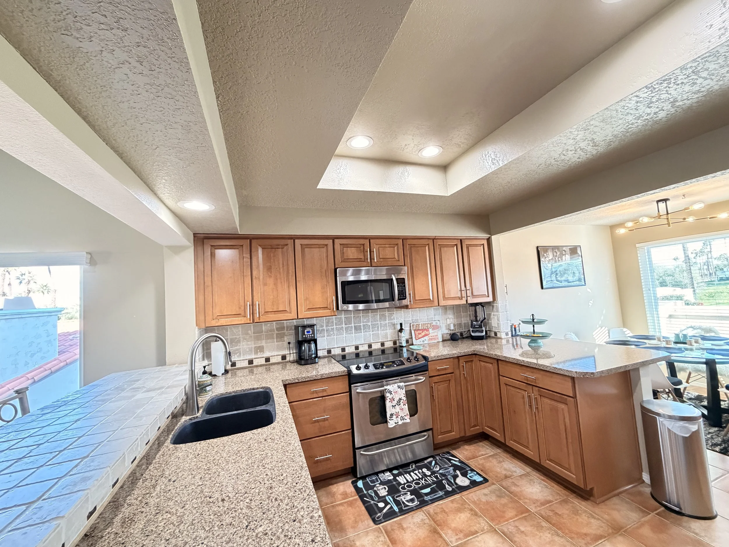 A kitchen with wooden cabinets, granite countertops, a stove, a microwave, and a sink. Recessed lighting and a window are visible above the counter, and a patterned rug is on the tiled floor.