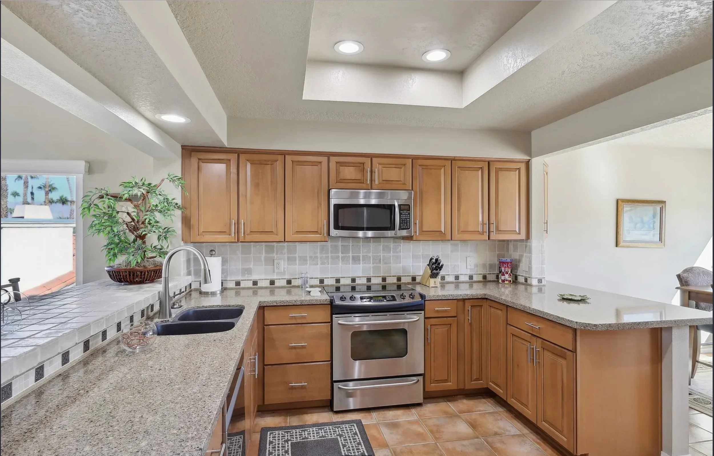 Kitchen with wooden cabinets, stainless steel microwave, oven, granite countertops, tiled backsplash, and a window with a view of palm trees outside.