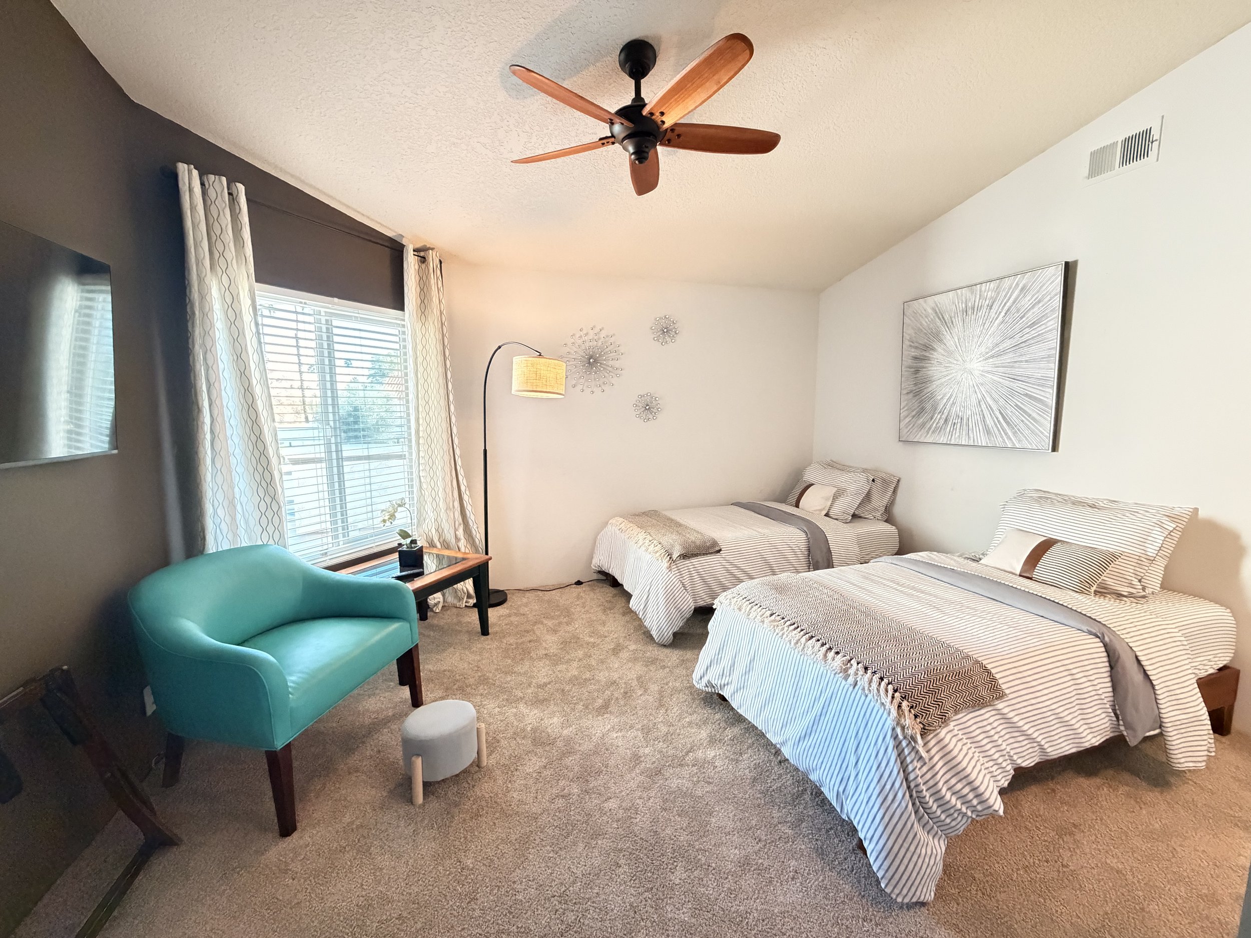 A bedroom with two twin beds with striped bedding, a teal armchair, a small wooden side table with a potted plant, a standing lamp, and wall art, with a window covered by white curtains and a ceiling fan.