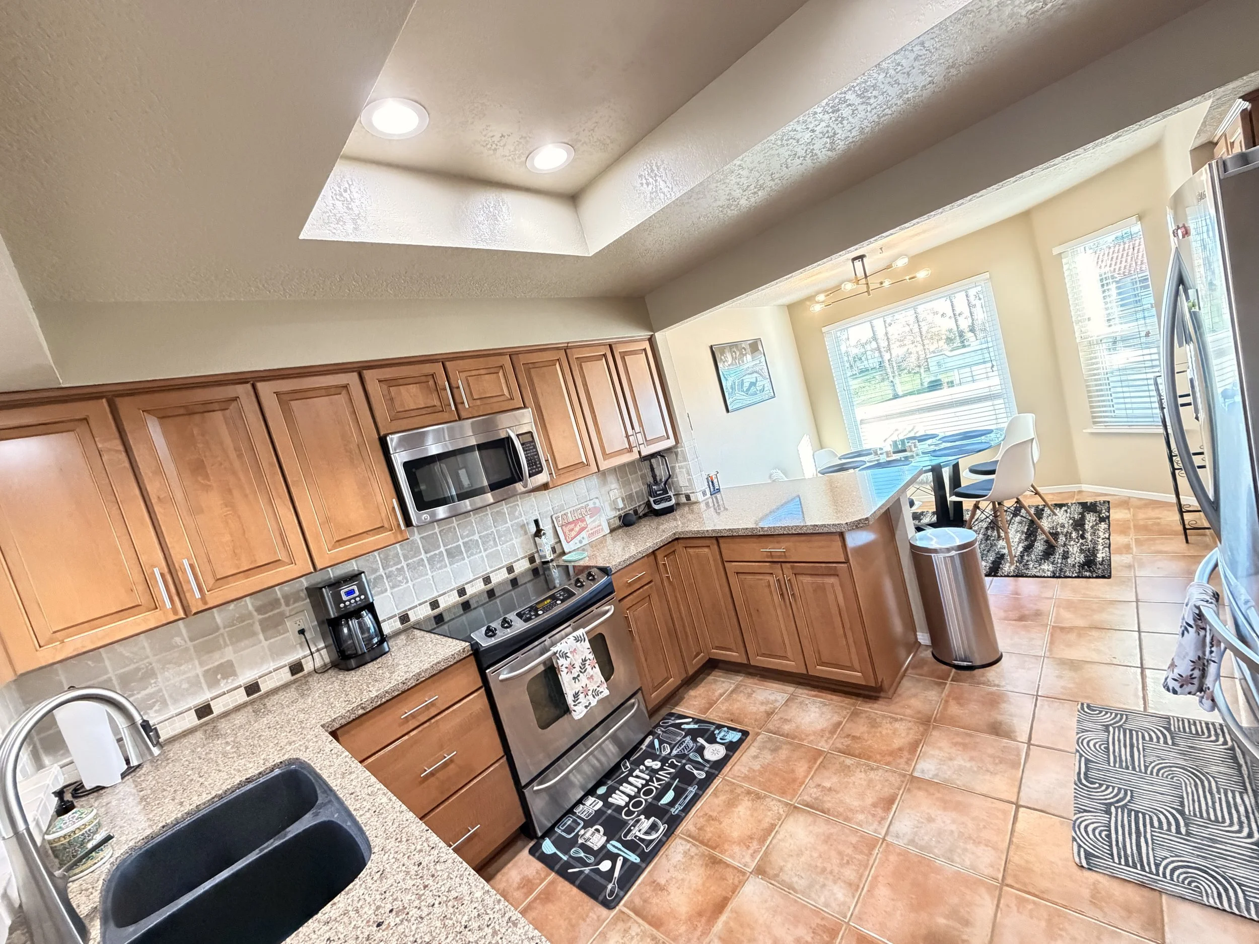 A modern kitchen with wooden cabinets, stainless steel appliances, a black sink, and a bright dining area with large windows and a round dining table with chairs.
