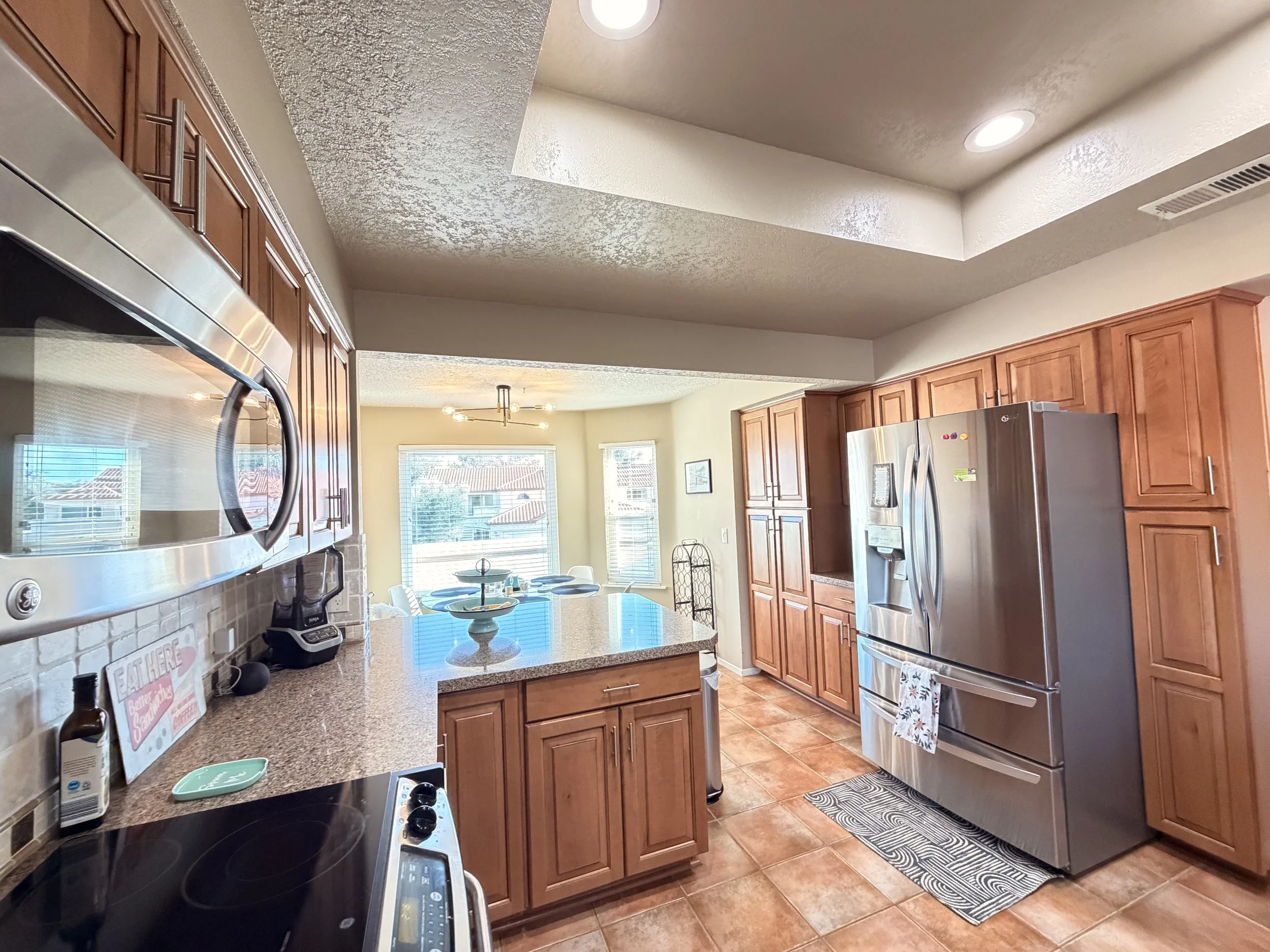 Kitchen with wooden cabinets, stainless steel refrigerator, microwave, black stove, countertops, and a dining table with chairs in front of large windows showing rooftops.