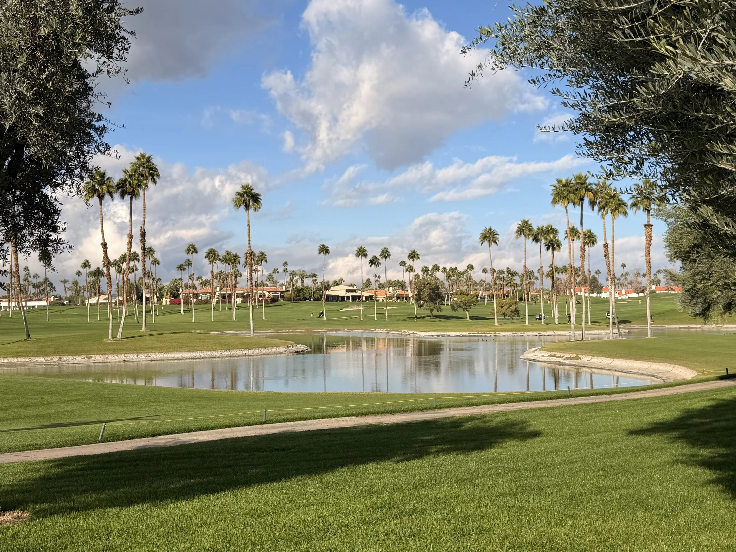 A golf course with lush green grass, several palm trees, and a small pond, under a partly cloudy sky.