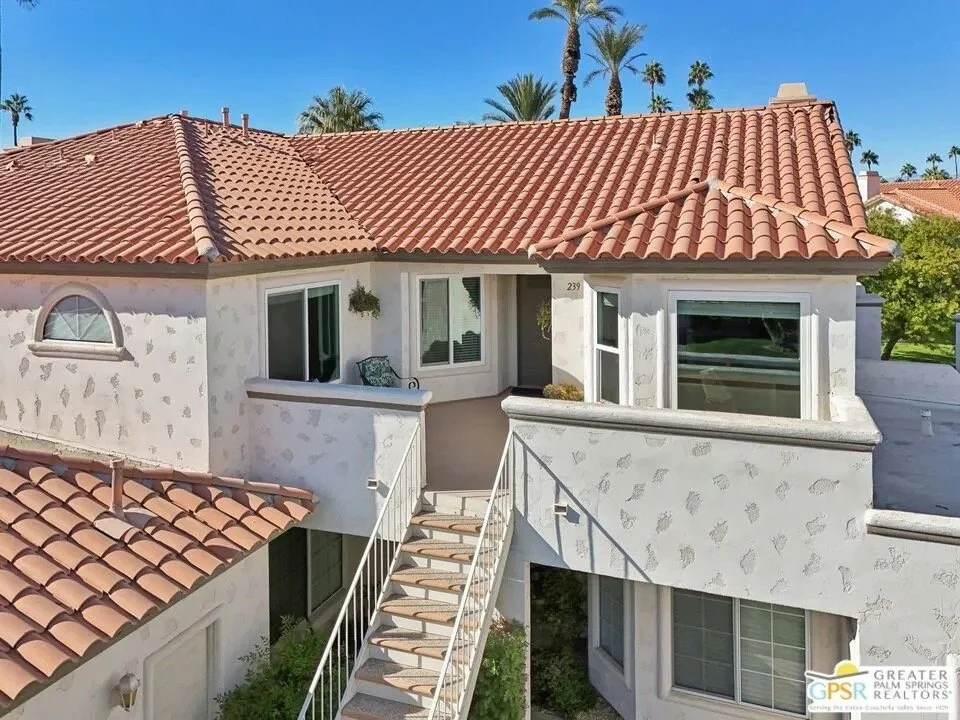 Front view of a white stucco house with a red tile roof and a small balcony, staircase, and large windows, with palm trees in the background.