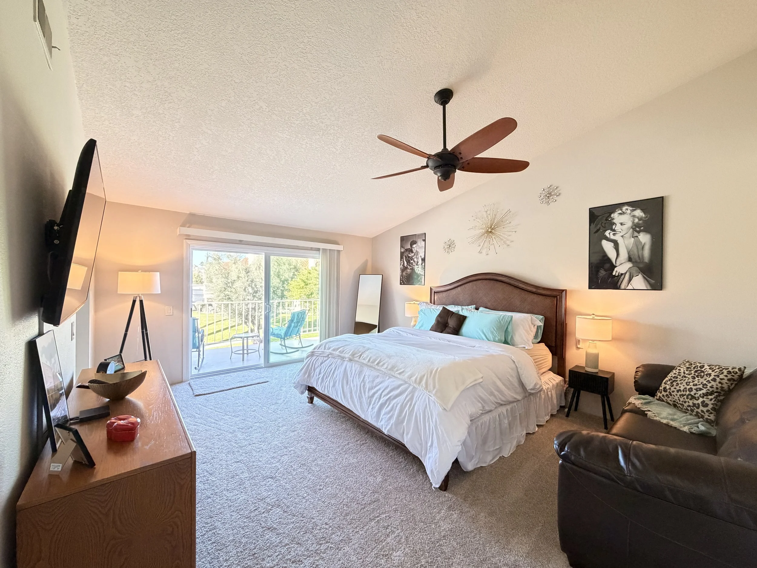 A bedroom with a bed on the right side, decorative wall art, and a sliding glass door leading to a balcony with outdoor seating.