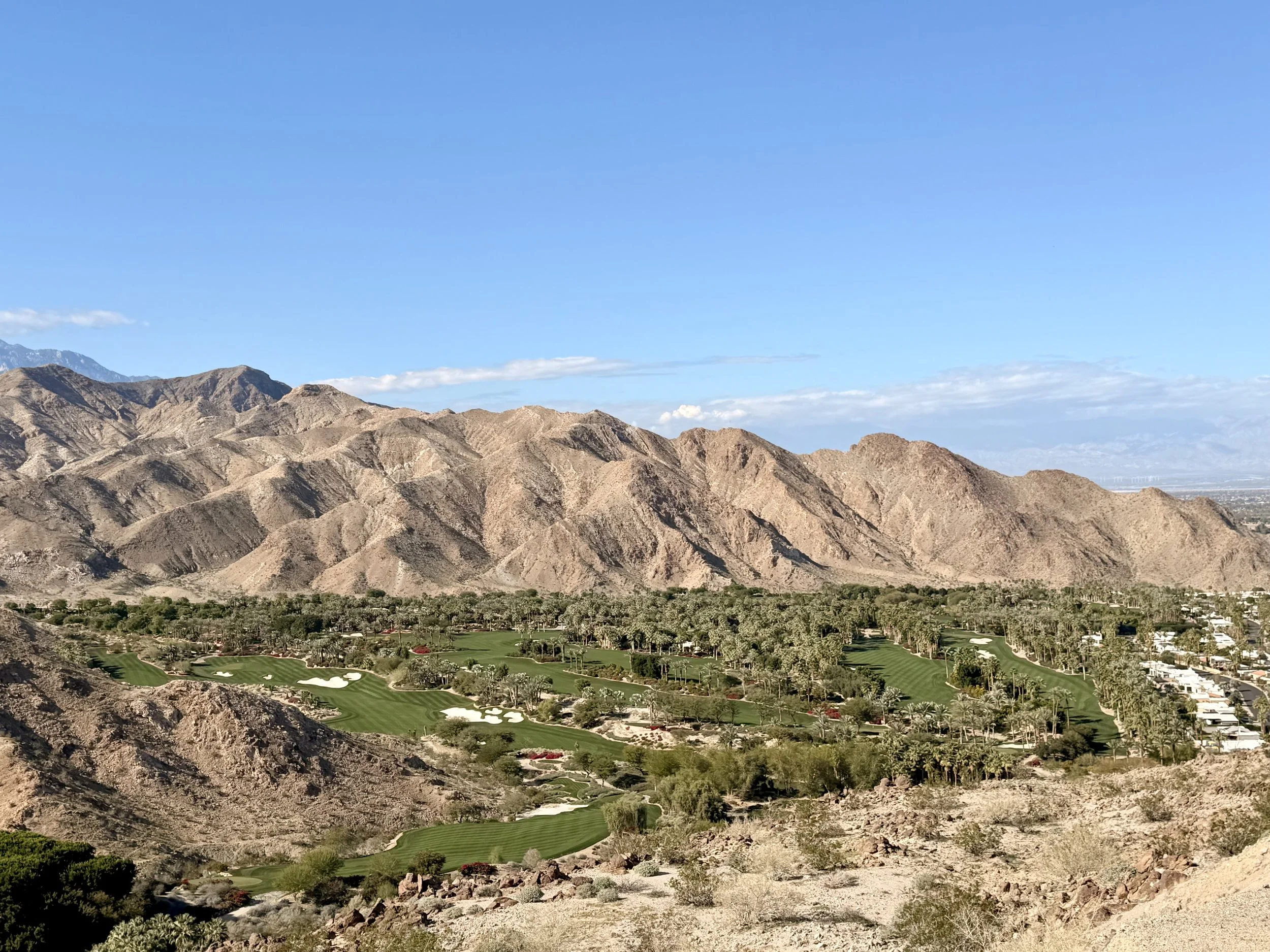 A desert mountain range with a golf course at the base featuring green fairways, sand traps, and scattered trees, with a clear blue sky overhead.