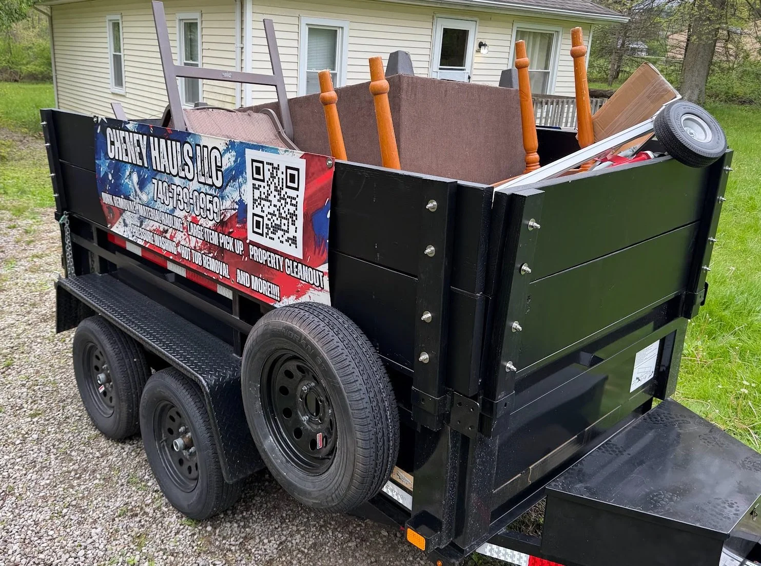 Black utility trailer filled with furniture and household items, parked on gravel in front of a house with beige siding and greenery.