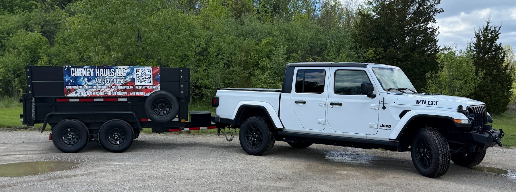 White Jeep Gladiator pickup truck pulling a black flatbed trailer with a sign for Cheney Hauls LLC, set against a green wooded background and a partly cloudy sky.