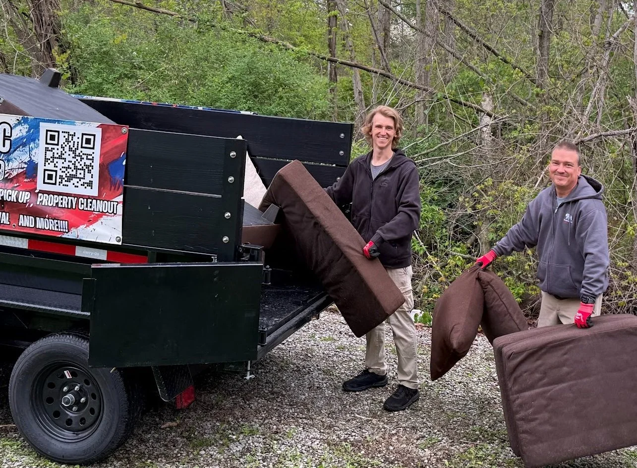 Two men in casual clothing and gloves loading furniture into a truck outdoors near trees and greenery.
