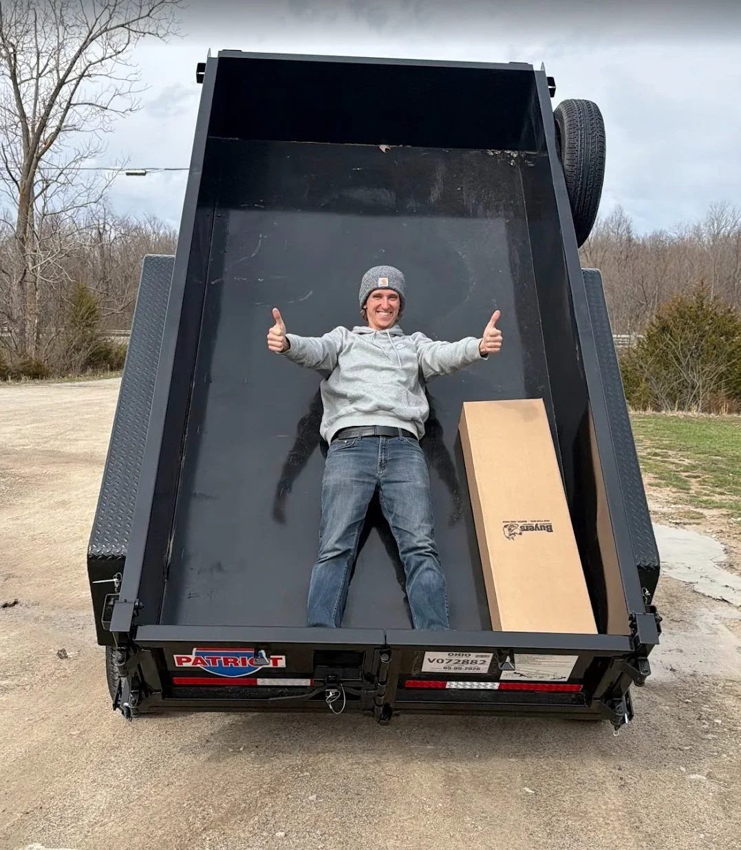 Person lying in a black utility trailer, smiling and giving two thumbs up, outdoors with bare trees and cloudy sky in the background.