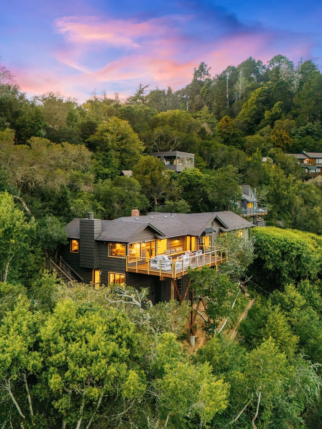 A house built on a hillside surrounded by dense green trees with a large deck illuminated by interior lights, under a pink and blue sunset sky.