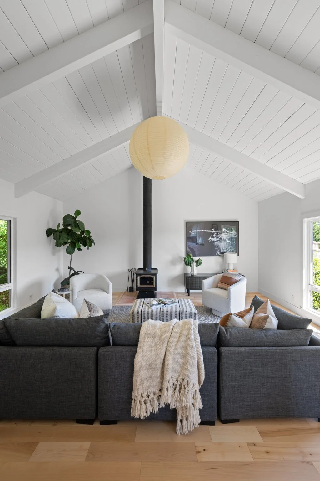 Living room with a vaulted white ceiling, a black wood stove with a chimney pipe, a large gray sofa with white and brown pillows, two white chairs, a striped ottoman, a potted plant, a side table with a lamp, a framed wall art, and large windows prov