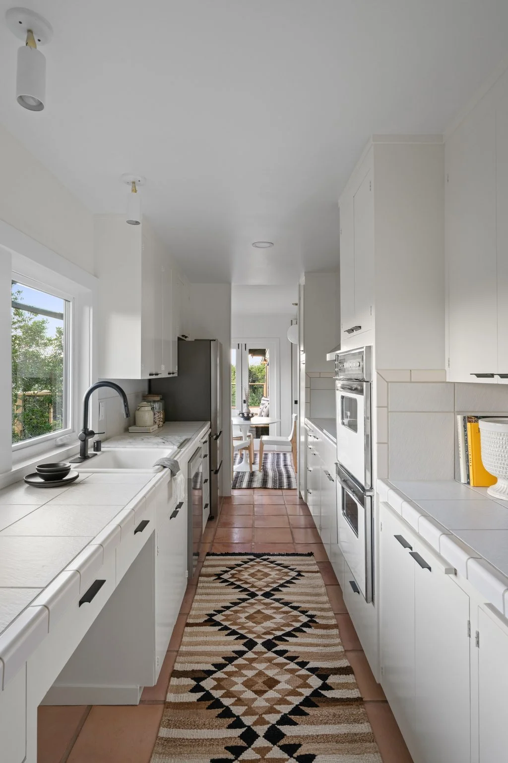 A bright, white kitchen with tiled countertops, a black faucet, and white cabinets. There is a window with greenery outside, and a decorative rug on the terracotta tile floor, leading to a dining area with chairs and a table.