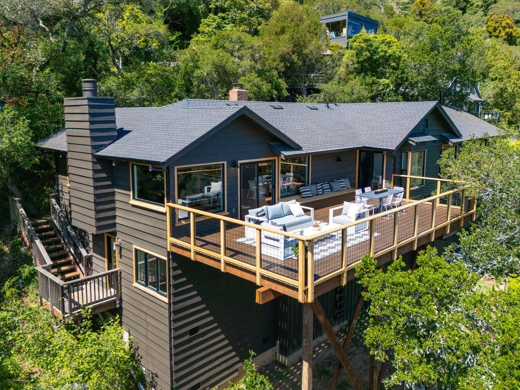 A house with a large wooden deck surrounded by trees, with outdoor furniture including sofas and chairs on the deck.