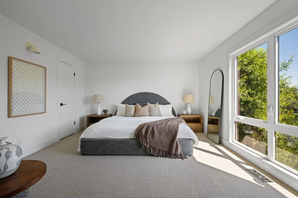 Bright, modern bedroom with large window showing green trees outside, bed with gray headboard, white bedding, beige and pink pillows, brown throw blanket, two wooden nightstands with lamps, a tall mirror, and framed artwork on the wall.