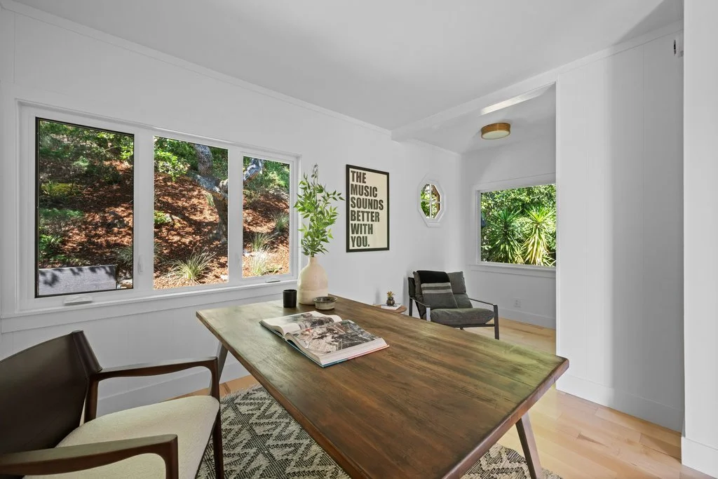 Bright, modern living room with large windows showing outdoor greenery, a wooden dining table with a plant and magazine, and minimalistic decor.