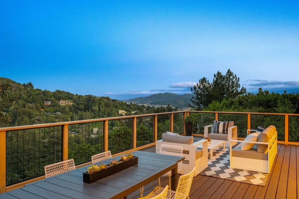 View of a spacious outdoor balcony with modern white sofa and chairs, a dining table with chairs, overlooking green hills and a blue sky.