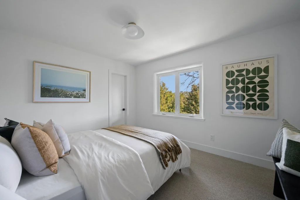 A bright bedroom with a bed covered in white bedding and throw pillows, a window showing trees outside, and framed artwork on white walls.