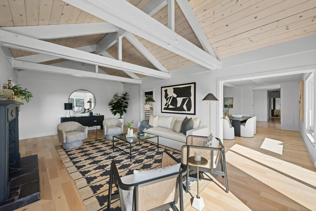 Living room with white walls, a wood plank ceiling with white exposed beams, and natural wood flooring. It features a white sofa, two patterned armchairs, a black sideboard with a round mirror above, a black and beige patterned rug, and a glass coffee table. In the background, there is an adjoining room with a dining table and chairs.