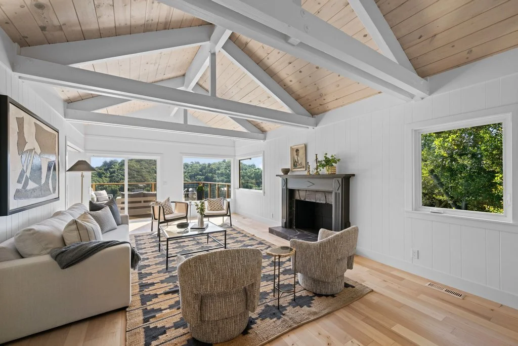 Bright living room with white walls and wooden ceiling beams, large windows, a fireplace, and modern furniture including a sofa, armchairs, and a coffee table.