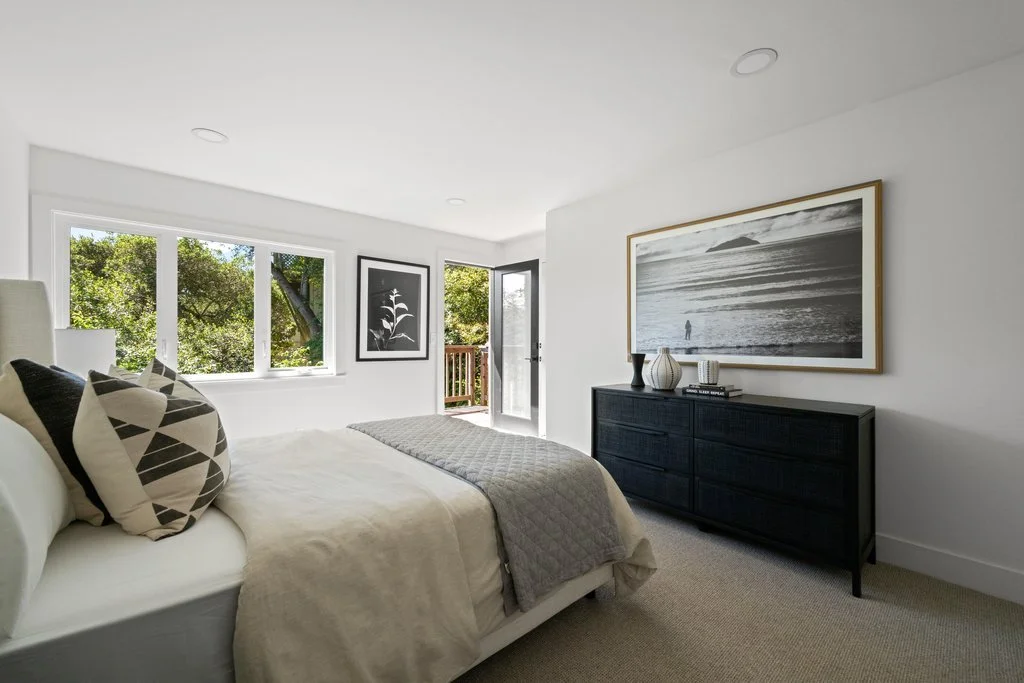 Bright bedroom with large window, white walls, decorated with framed artwork, black dresser with vases, and a bed with patterned pillows.