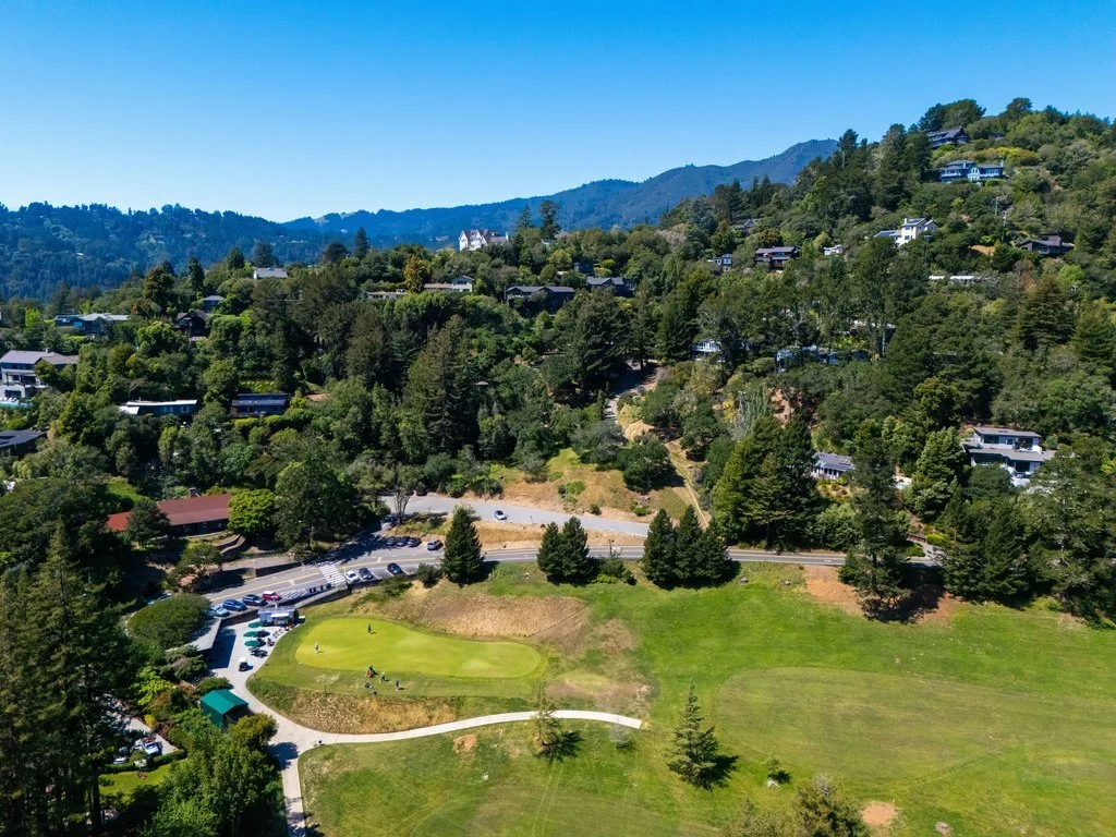 Aerial view of a golf course surrounded by lush green trees and houses on a hillside, with a parking lot and a small building near the golf course.
