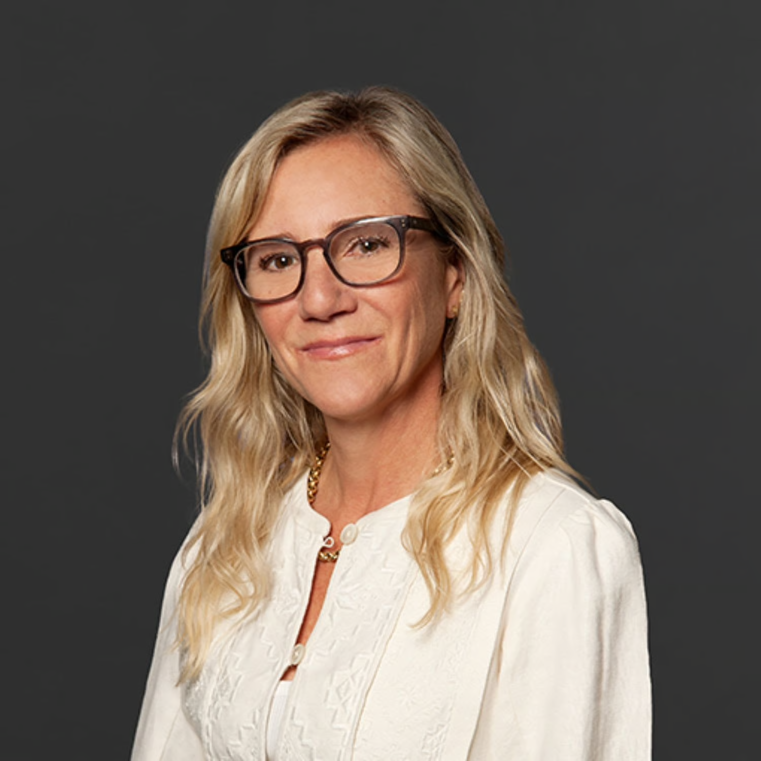 Portrait of a woman with blonde hair, wearing glasses, a white top, and gold jewelry, against a dark background.