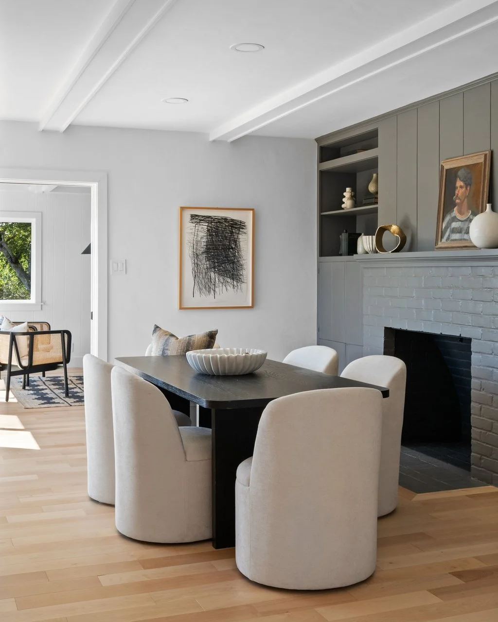 A dining room with a dark wood table surrounded by five white upholstered chairs, a white painted brick fireplace, a built-in bookshelf with decor, and wall art, with a bright window showing outdoor greenery.