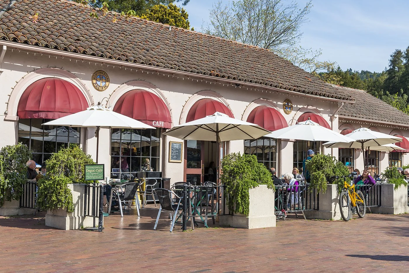 Outdoor seating area of a cafe with umbrellas, tables, and chairs. The cafe has a white exterior with red awnings and potted plants. People are sitting and enjoying their time outside.