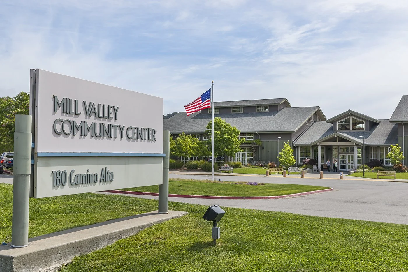 Exterior of Mill Valley Community Center with a sign in the foreground and a building with gray roofing and windows in the background. An American flag is flying on a flagpole near the entrance, and there are trees, benches, and people outside.