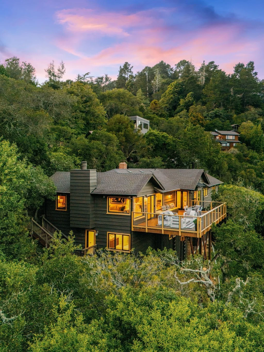 A house with warm yellow lights on a hillside surrounded by dense green trees and a colorful sky at dusk.